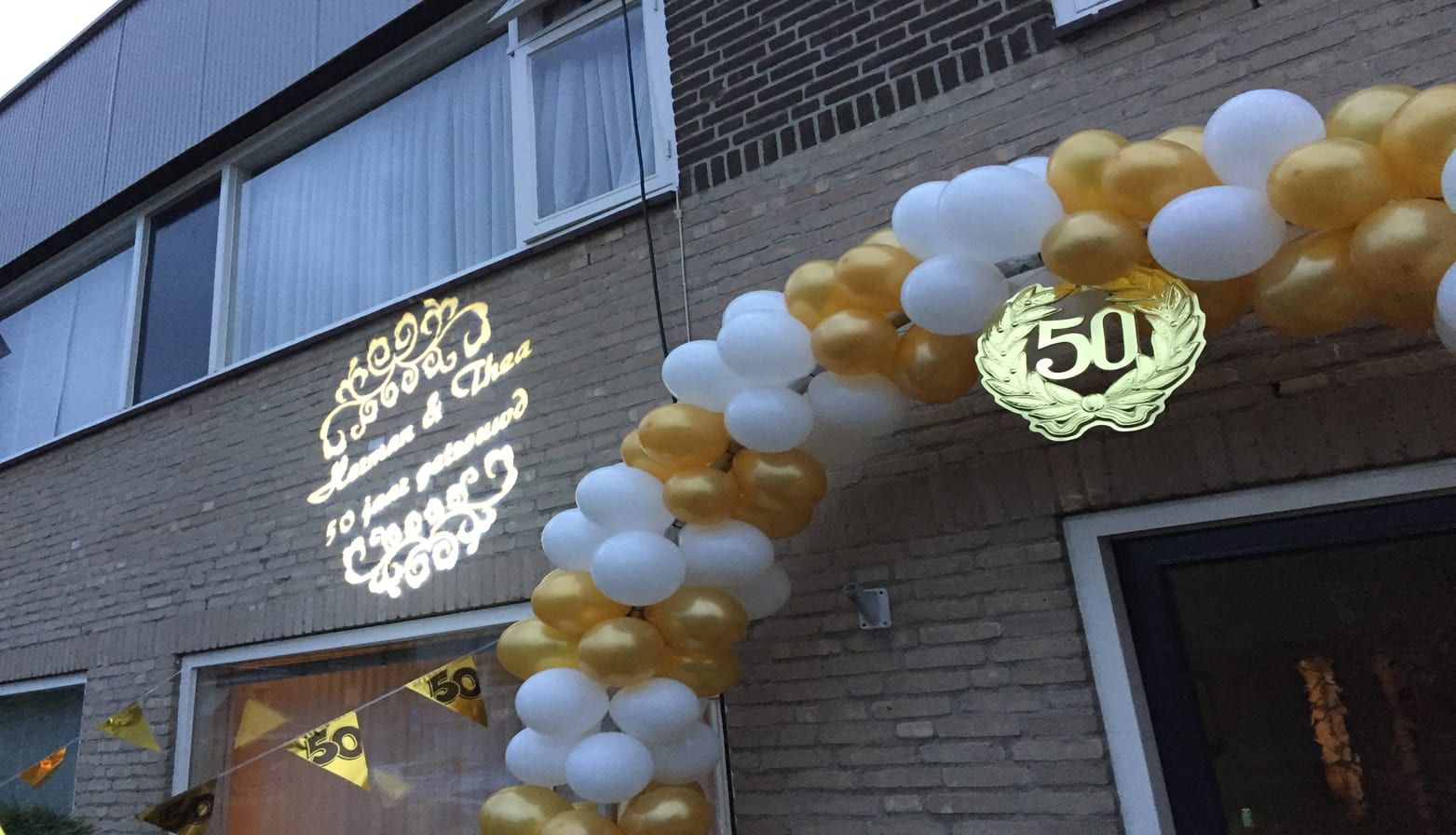 Gouden gobo-projecties voor een 50-jarig huwelijk op een gevel, naast een ballonnenboog
