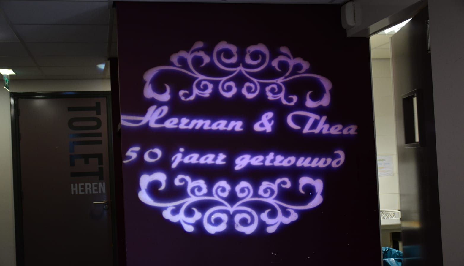 Sierlijke gobo met de tekst Herman & Thea 50 jaar getrouwd in paars licht op een wand