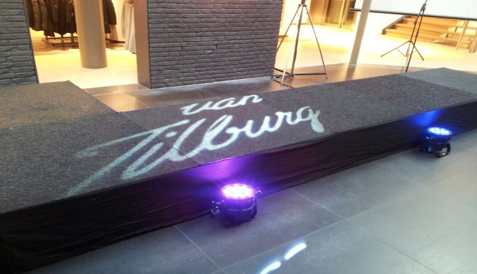 Logo van Van Tilburg als gobo geprojecteerd op een catwalk, tussen paarse spots